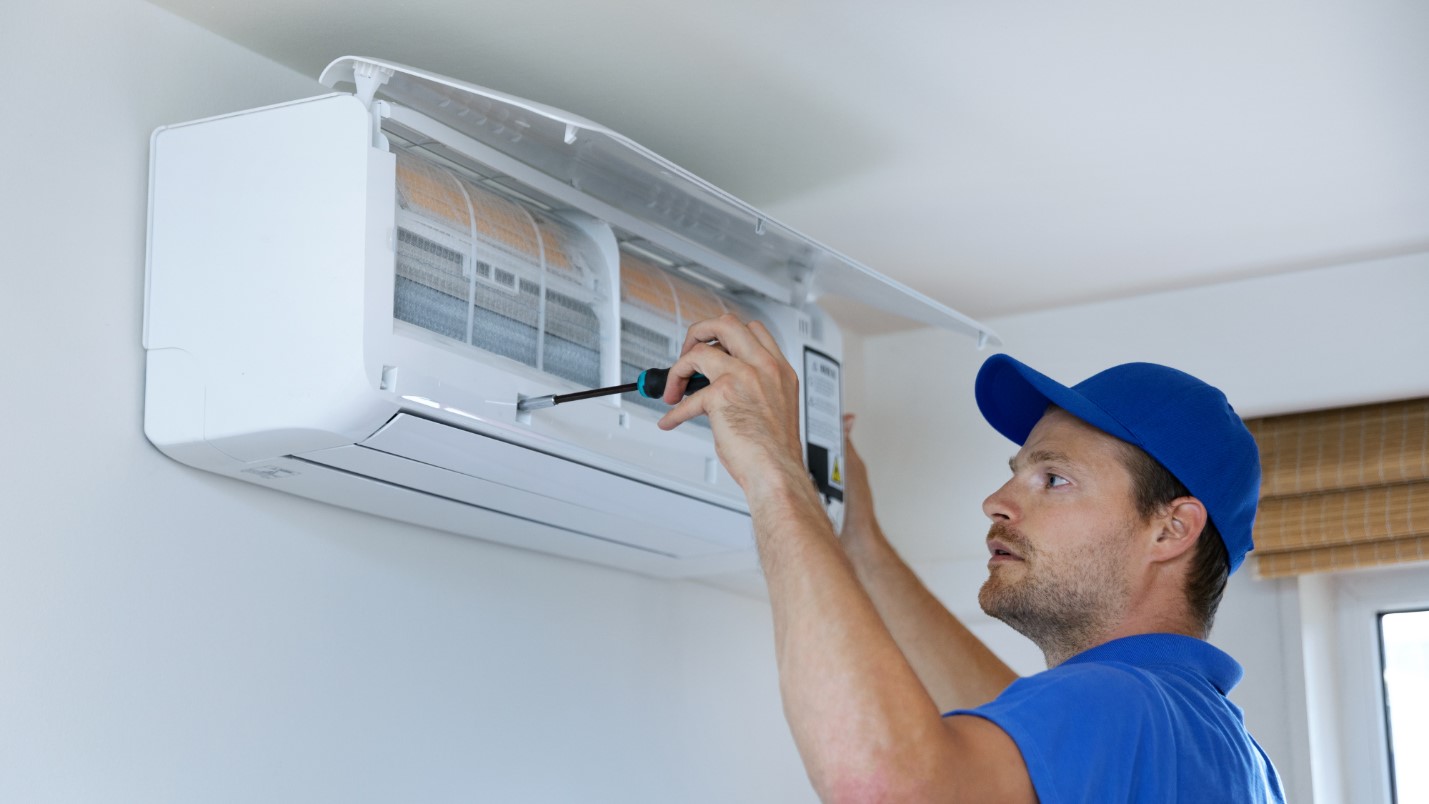 A man in a blue hat uses a screwdriver to adjust a component on an AC unit.