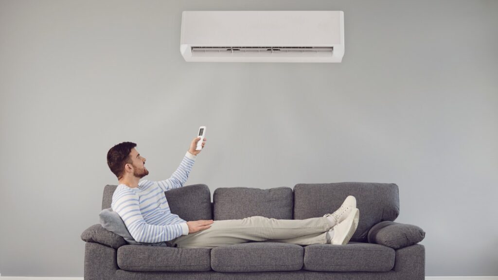 A man on a couch uses a remote to adjust an AC placed on the wall above him.