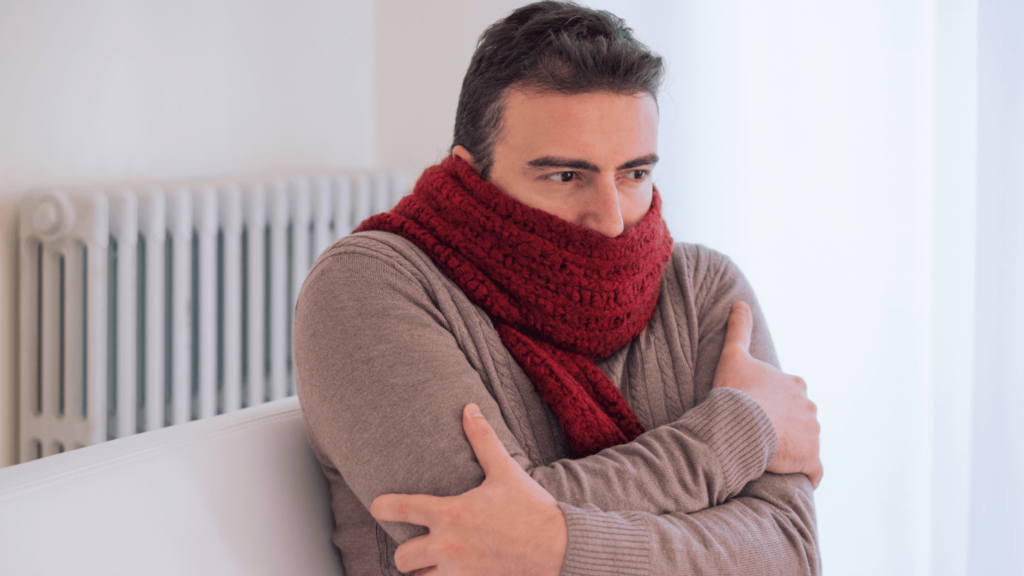 A man wearing a scarf sits on a chair in a living room, he is holding his arms to keep warm.